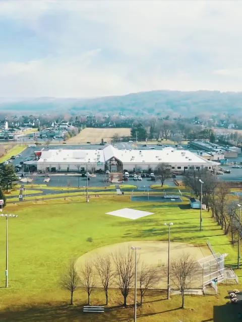 Aerial view of the gym tiME sports complex surrounded by green fields and parking lots, with the facility's logo overlaid on the image.