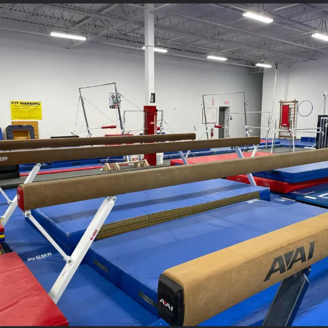 Spacious gymnastics gym featuring balance beams, mats, and bars equipment with safety padding in a well-lit facility.