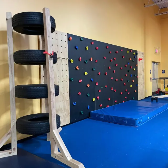 A gymnastics training area with a colorful climbing wall and a tire setup next to a blue practice mat.