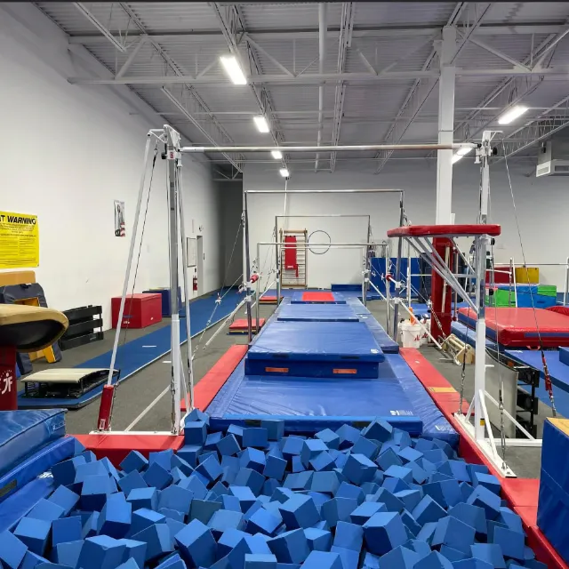 A well-equipped gymnastics facility with uneven bars over a foam pit, a vault table, and balance beams in the background, ready for training.