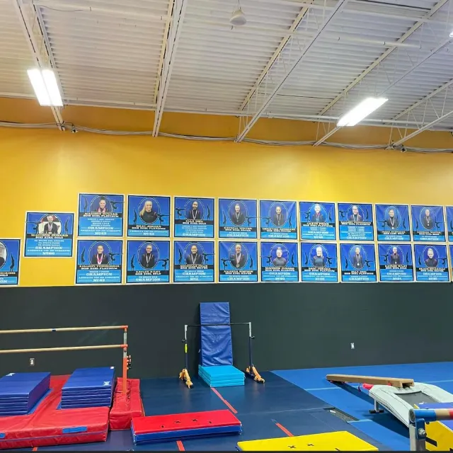 Interior of a gymnastics hall with a 'Wall of Champions' featuring athletes' portraits and achievements, along with training equipment such as balance beams and mats.