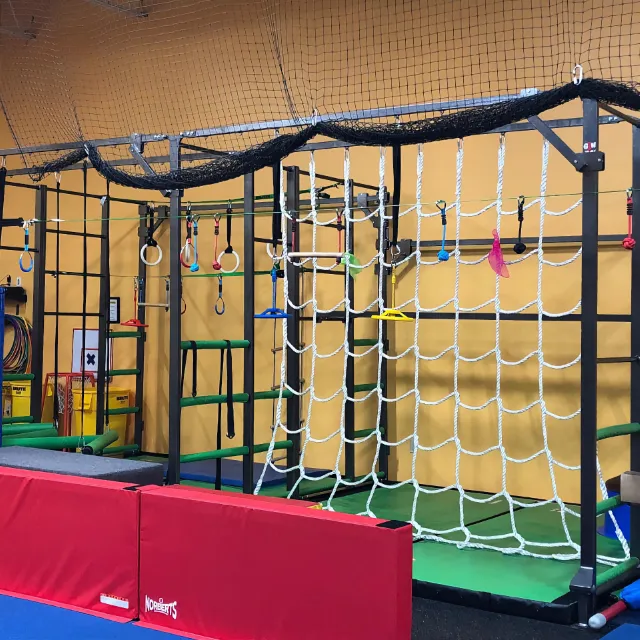 An indoor gymnastics obstacle course with climbing nets, rings, and bars, featuring colorful equipment against a yellow wall, designed for children's agility and strength training.