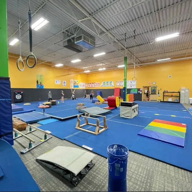 A wide view of a gymnastics training facility featuring various equipment such as mats, balance beams, and rings with people practicing in the background, under bright lighting.