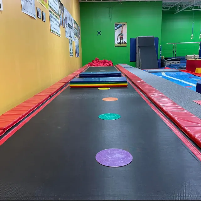 Long tumbling track in a gymnastics gym with colorful circular spots for jump training, flanked by red protective padding.