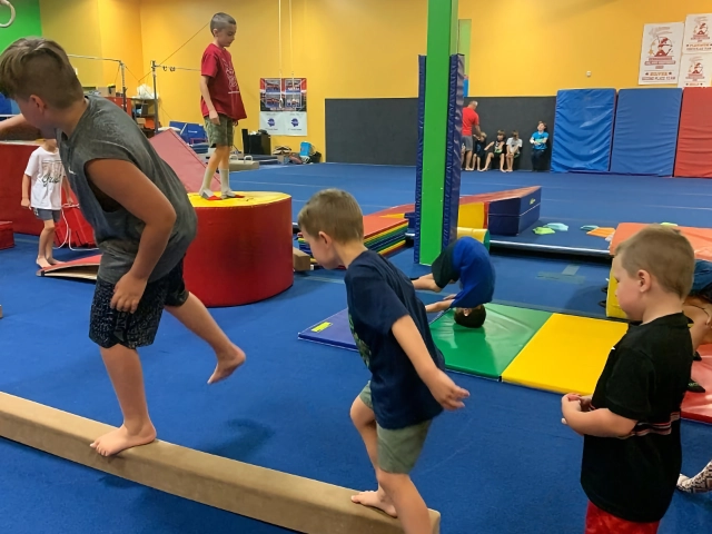 Young boys practicing walking on a balance beam in a gymnastics class with colorful equipment around them.