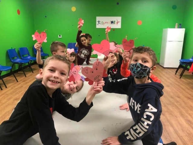 A group of children showing off their handmade heart-shaped crafts, smiling and enjoying their time in a room with green walls.