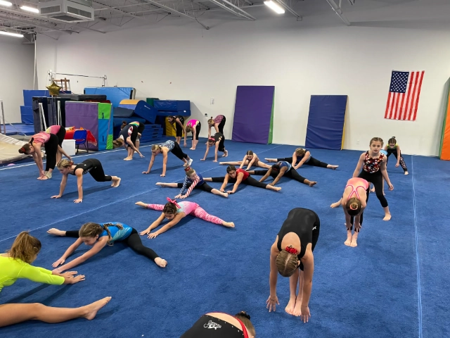 A group of young gymnastics students engaging in flexibility training on mats in a spacious gym, with an American flag in the background.