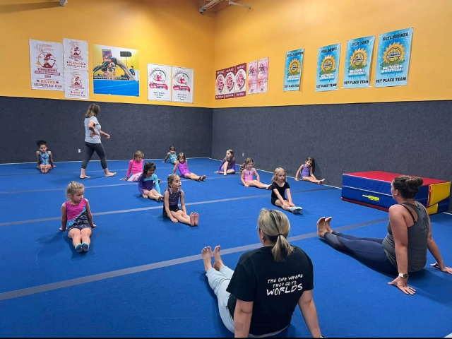 Children sitting on blue mats following their instructor's stretching routine in a gymnastics class with achievement banners on the wall.