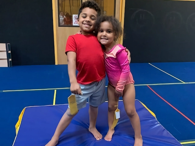 Two young siblings hugging and smiling on a gymnastics mat, showing affection and happiness in the gym.