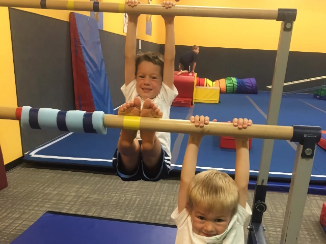 Two young boys with smiling faces hanging from gymnastics equipment, enjoying their class in a colorful gym.