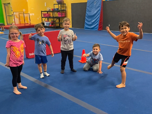 A group of cheerful children posing with thumbs up in a gymnastics class, showing excitement and readiness.