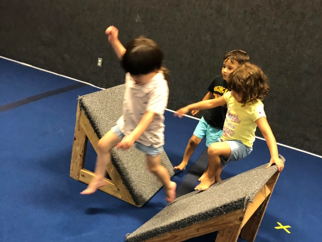 Three children energetically jumping over and playing with foam blocks in a gymnastics training room.