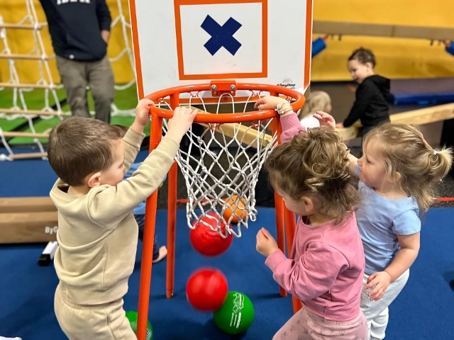 Toddlers excitedly playing with a toy basketball hoop in a play gym, with an adult supervising in the background.