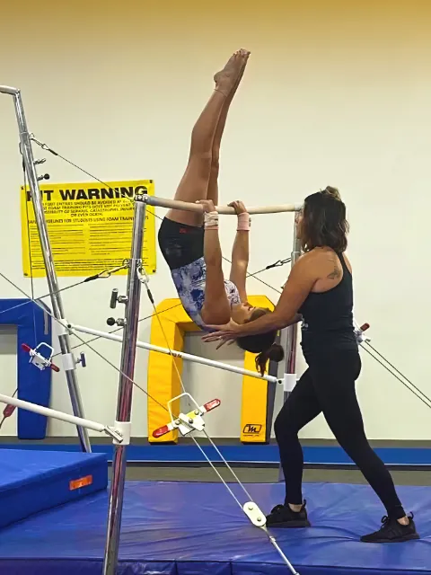 A young gymnast in an inverted position on the uneven bars while being assisted by her coach during training.