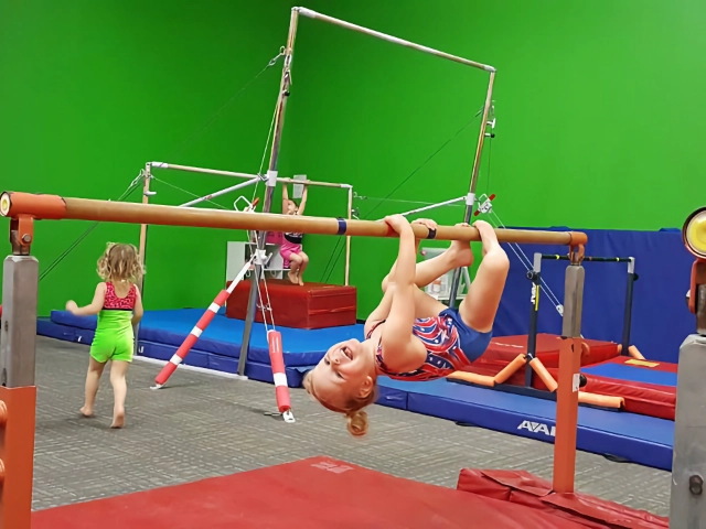 A joyful girl hanging upside down on the uneven bars in a gymnastics gym with green walls and another child in the background.