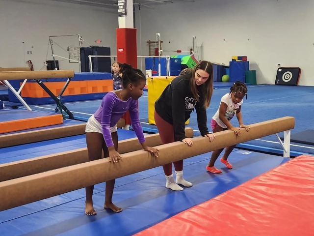 A gymnastics coach instructing two young girls on the balance beam in a gym, with other children practicing in the background.