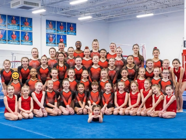 A large group of gymnasts in red leotards posing for a team photo in a well-equipped gymnastics facility.