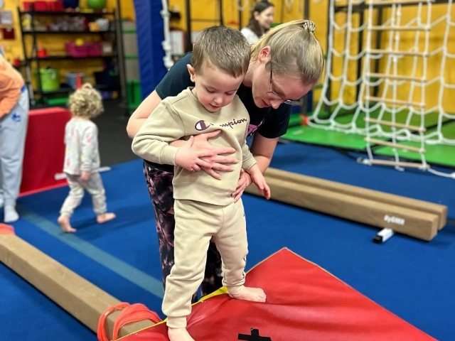 A toddler receiving assistance from a gymnastics instructor while stepping onto a red mat in a gym, with other children playing in the background.