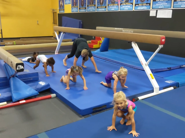 Children in a gymnastics class performing crawling exercises on blue mats, with balance beams and gymnastics equipment in the background.