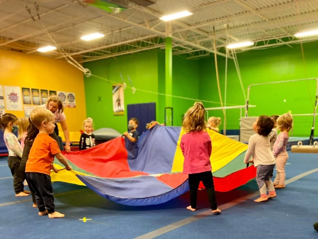 A group of children playing with a large, colorful parachute in a vibrant gymnastics gym.