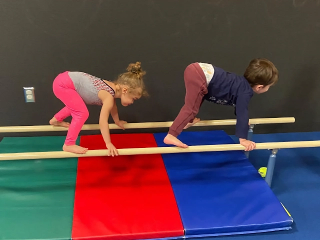Two young children practicing their balance on a low beam in a gymnastics class, with colorful mats on the floor for safety.