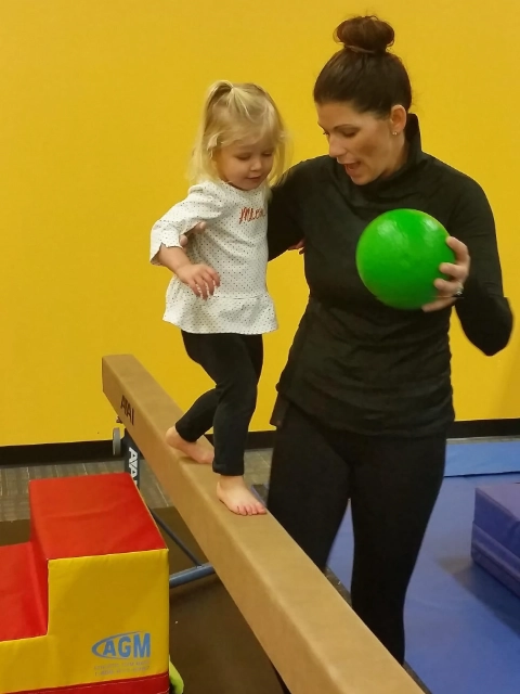 A young toddler with her coach practicing walking on a balance beam in a gymnastics class, with the coach holding a green ball.