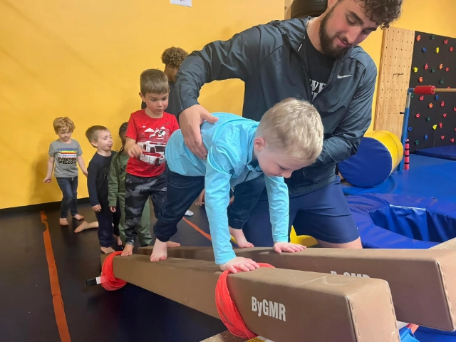 A gymnastics coach assisting a young boy on the balance beam while other children wait their turn in the background.