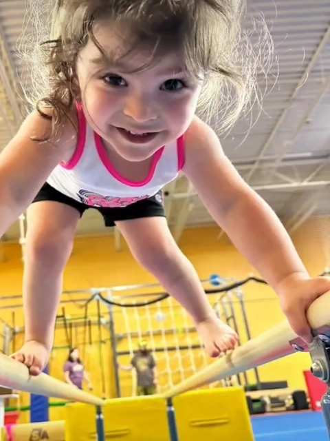 Close-up of a smiling young girl hanging from a gymnastics bar, with gym equipment in the background.