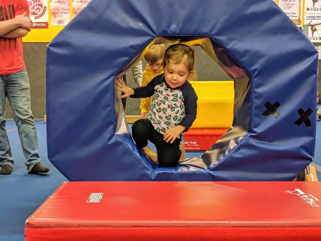 A toddler emerging from a play tunnel at a gymnastics class with other children in the background.