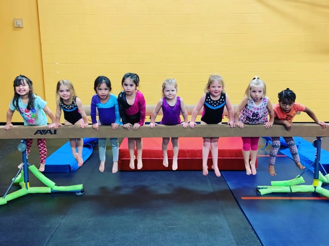 A line of young gymnasts hanging from a low balance beam with focused expressions, in a gym with yellow walls.