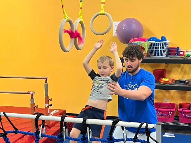 A young boy being coached on gymnastic rings training with the assistance of a male coach in a colorful gym.