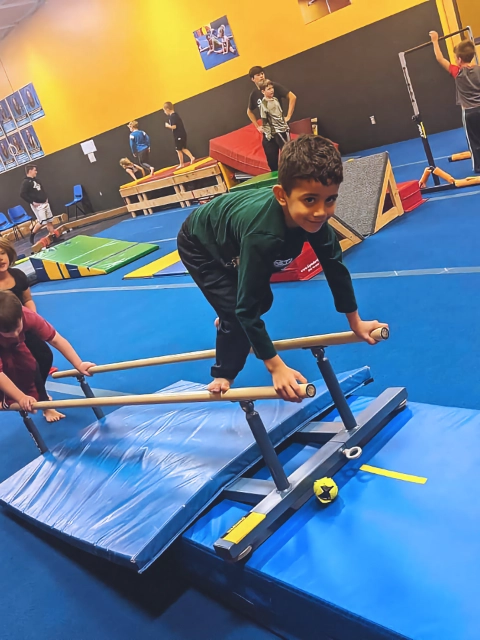 A young boy focusing on training with parallel bars in a gymnastics gym, with other children practicing in the background.