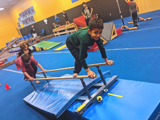 A young boy focusing on training with parallel bars in a gymnastics gym, with other children practicing in the background.