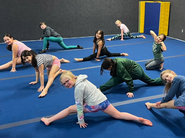 A diverse group of children smiling and stretching on a blue gym floor during a gymnastics class.