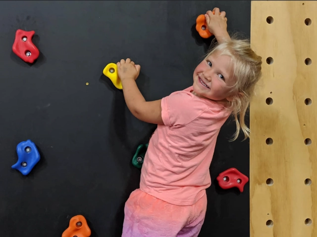 A smiling young girl enjoying climbing on a colorful indoor rock wall, looking at the camera.