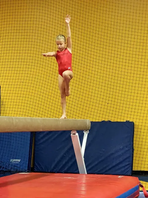 A young gymnast performing on a balance beam, confidently raising her arm in front of a yellow mesh background.