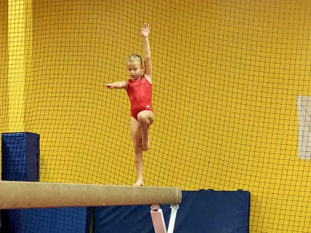 A young gymnast performing on a balance beam, confidently raising her arm in front of a yellow mesh background.