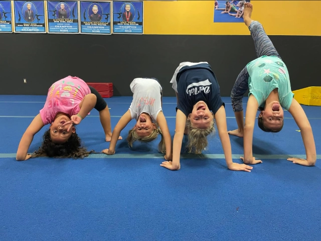 Four girls practicing backbends on a blue gym mat, displaying flexibility during gymnastics practice.