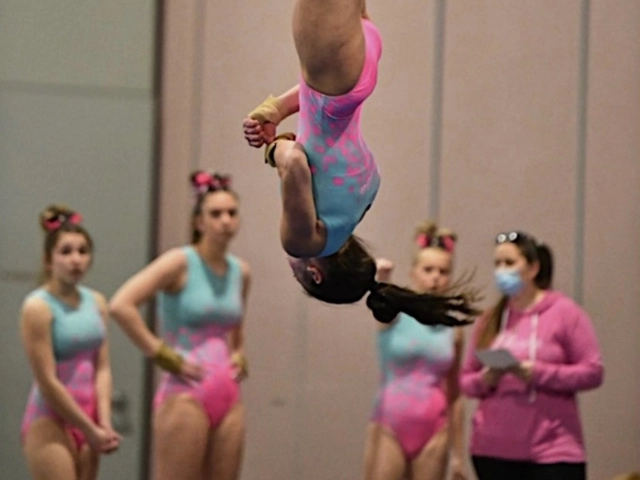 A gymnast captured mid-flip during a gymnastics competition, with judges and other competitors in the background.