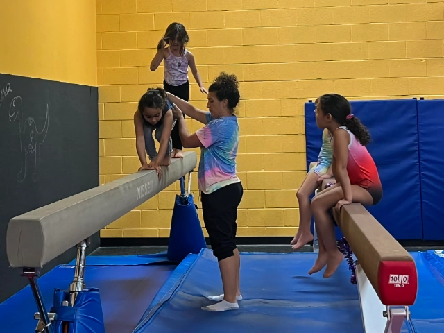 A gymnastics instructor is carefully assisting a young girl on the balance beam while others wait their turn in the gym.