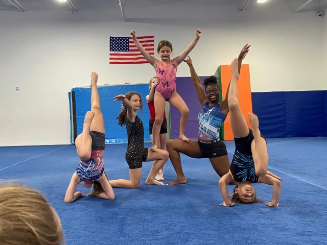 A joyful group of children posing in various gymnastics positions on the mat, with an American flag in the background, during their class.