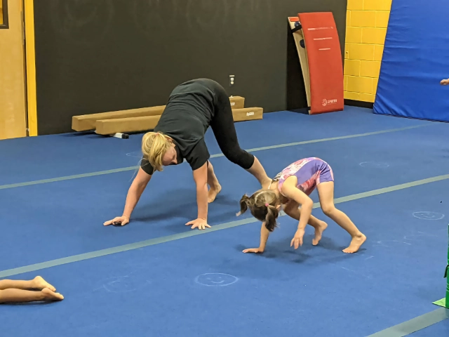 A gymnastics instructor guiding a young girl through a cartwheel practice on the gym floor.