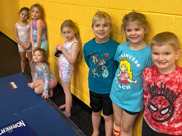 A lineup of kids with various expressions waiting for their gymnastics class against a bright yellow wall.