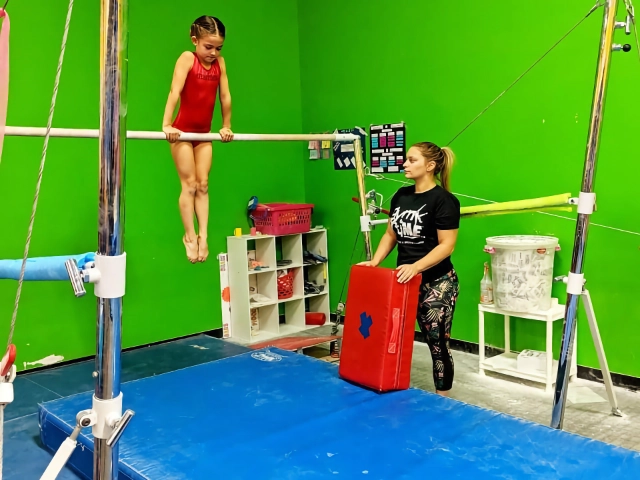 A young gymnast in a red leotard practicing on the uneven bars with her coach attentively watching, in a gym with bright green walls.